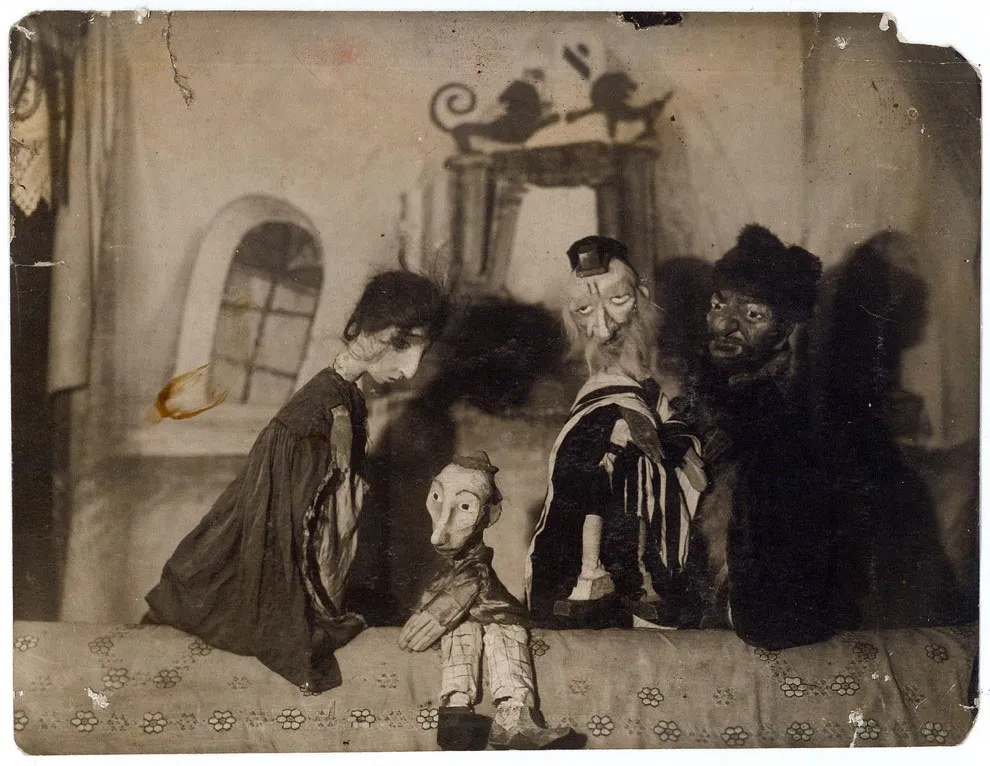 Four puppets stand gathered in the center of a stage displaying with a set of a small village.