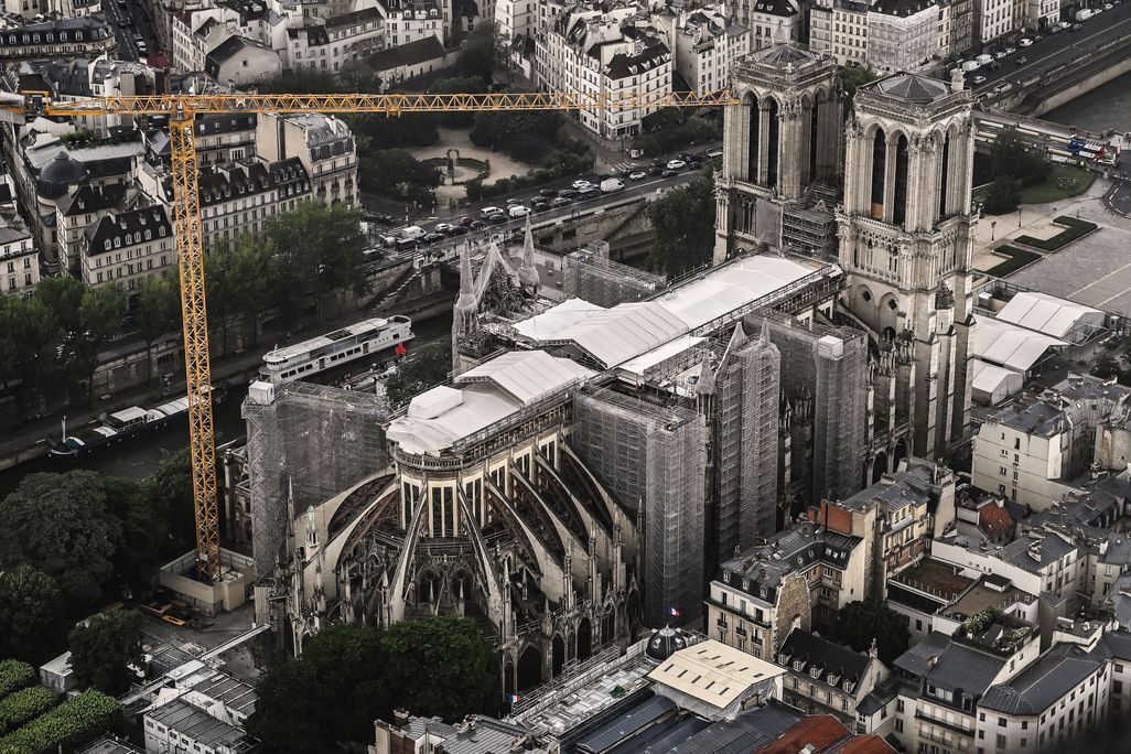 An aerial view of the exterior of the cathedral, its damaged roof mostly covered by white tarp. An enormous crane looms over the building