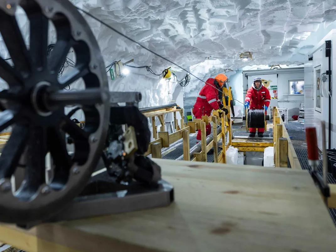 a metal wheel with cables leads from the foreground back to a pair of researchers in an ice cave