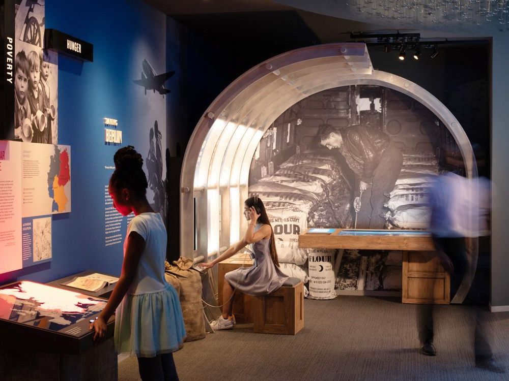 Two young people sit and stand in front of exhibits, reading the text; the blue walls are richly decorated with photographs, maps and information about post-war Europe