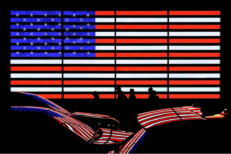 OPENER - An illuminated American flag display casts its colors onto passing cars, merging national symbolism with New York City’s constant flow of people and light.