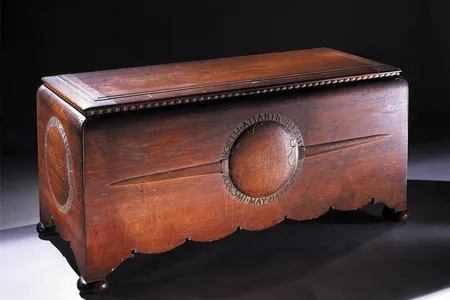 A large hand-carved teakwood chest with scalloped edges and four round feet. The chest appears to be stained a rich reddish brown. The front panel on the chest has a bas-relief globe pattern flanked by a pair of airplane wings.