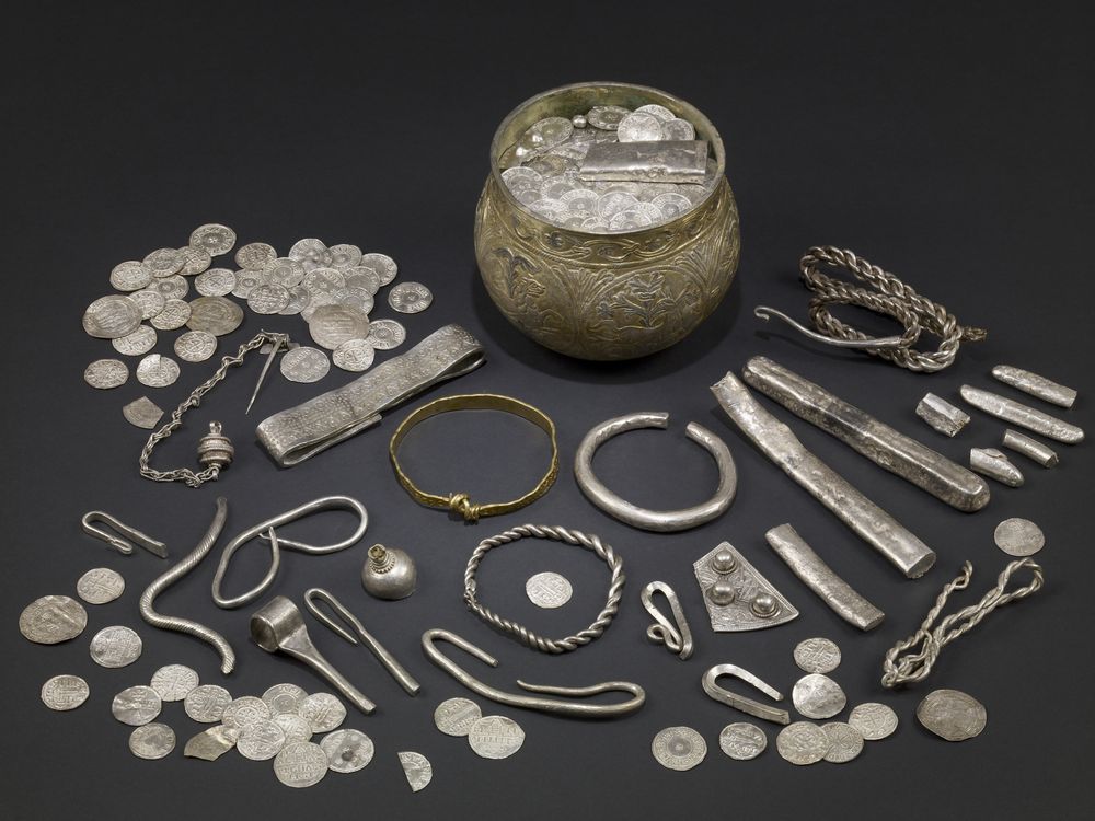 A display of silver Viking artifacts laid out on a table, including a wide mug-like cup, many coins, silver bars and what appear to be bracelets and other jewelry scattered around