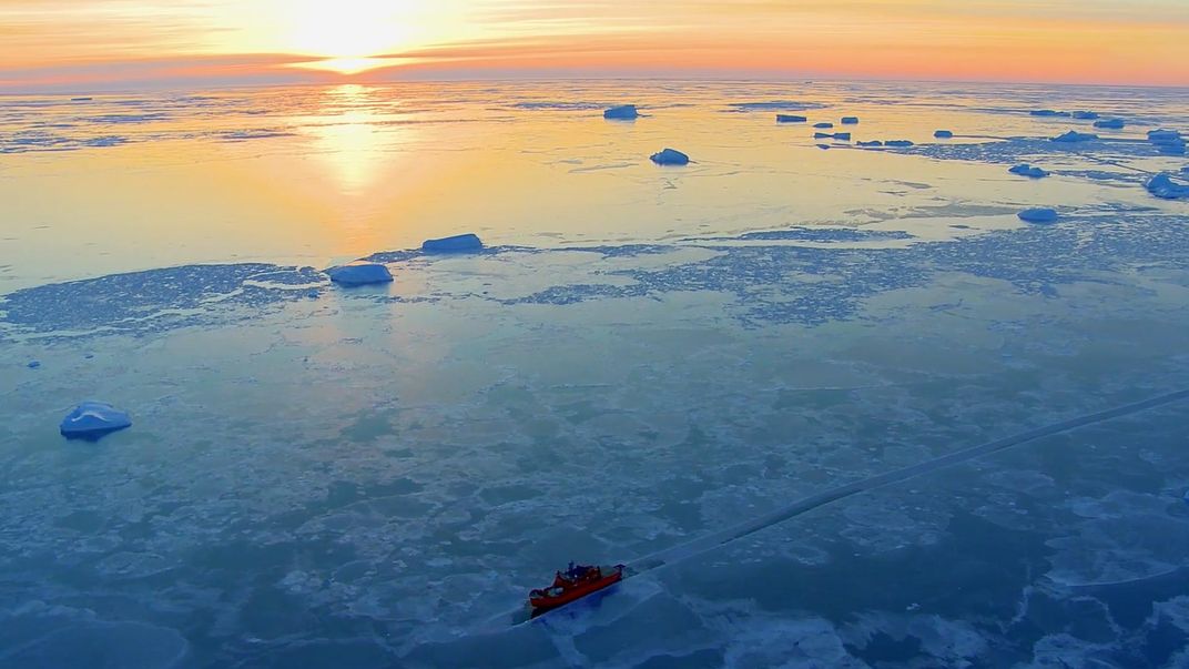 A ship sails through ice sheets on the ocean as the sun sets.