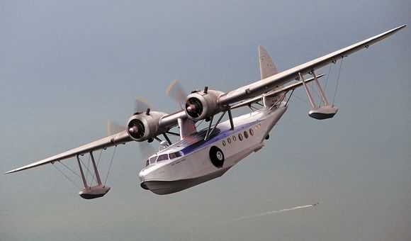 Silver twin engine flying boat over a vast ocean. A small boat leaves it's wake below.