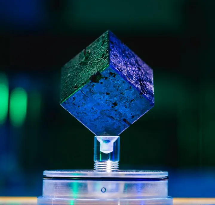 A blue and green image of a small metal cube resting on a display, with colored lights in the background