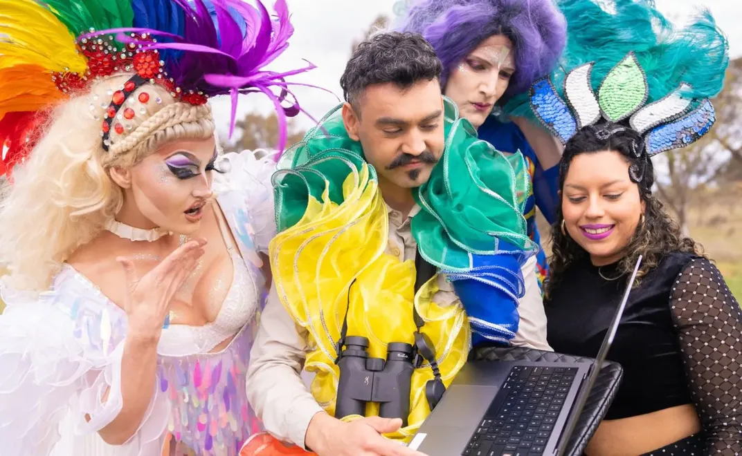 Weliton Menário Costa, wearing a boa, stands holding a laptop with binoculars around his neck; on the left is a drag queen, looking at the laptop, and two dancers, dressed in purple and black respectively, stand on the right, also looking at the screen.