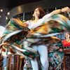 A person dances, turquoise and brown skirt flying and twisting around them, with a drummer and singer performing on the sides.