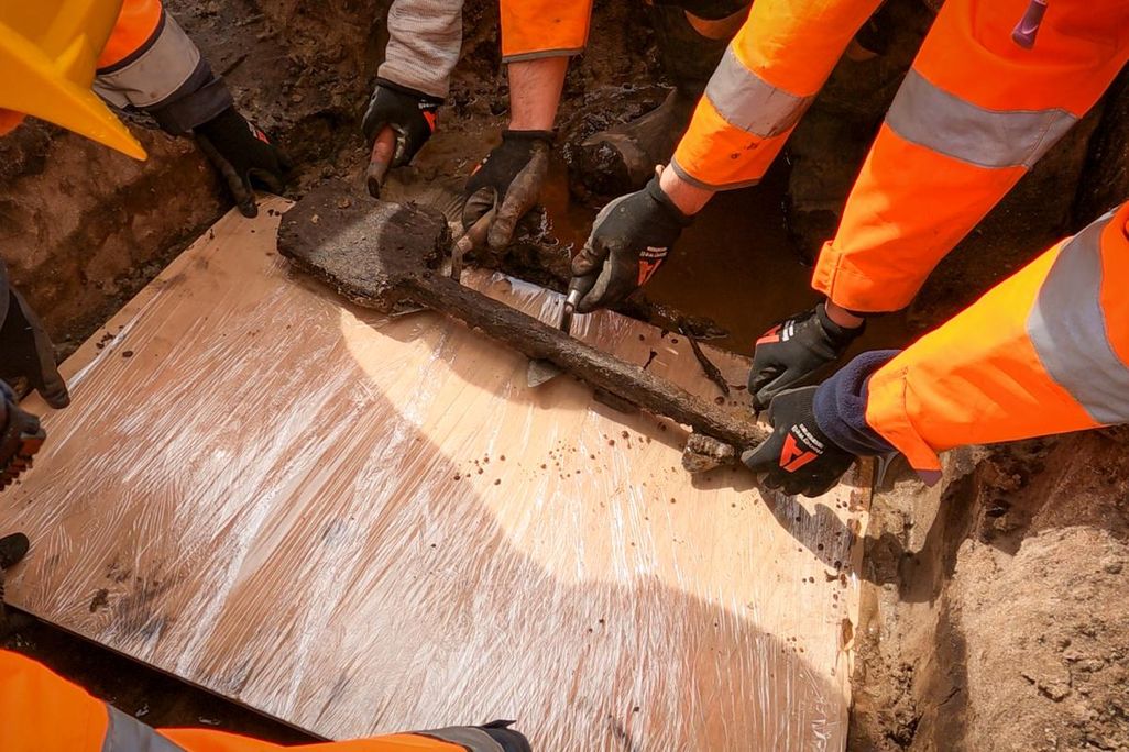 People wearing orange jackets lifting a wooden spade onto a piece of flat wood