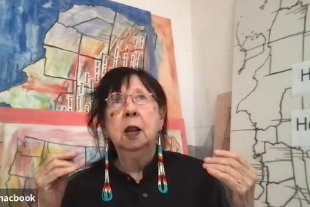 Still from a video of Jaune Quick-to-See Smith. Smith wears a black shirt with her hair pulled back, gold wire glasses, and long beaded earrings. She is sitting in front of works of art depicting maps of the United States, several in color and one in blac