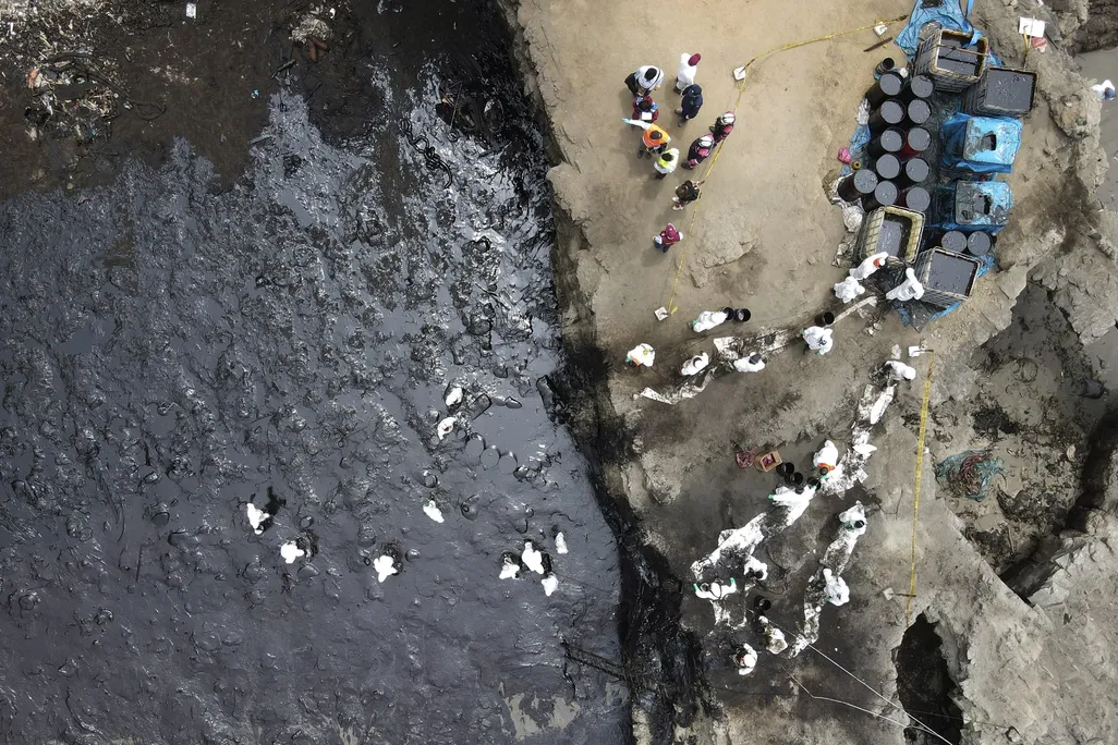 An aerial image of an oil spill off Peru's coast. The photo shows a contaminated beach smeared with black crude oil and workers in PPE trying to clean up the beach.