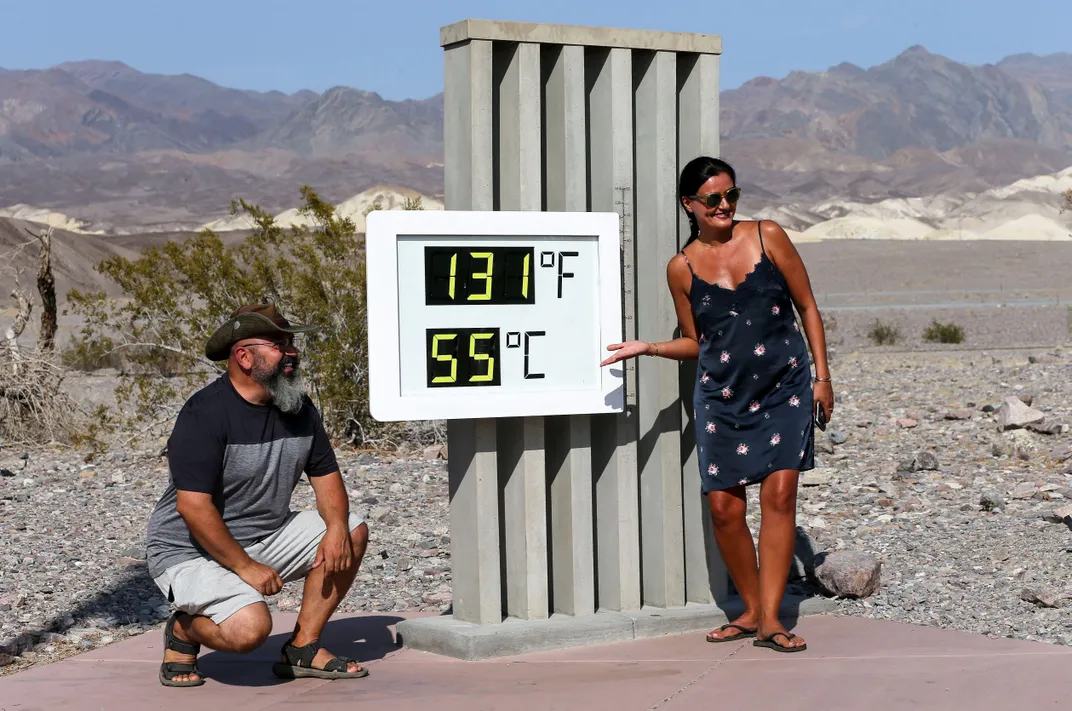 A man in a cowboy hat kneels and a woman in a sundress stands and gestures at a large digital thermometer, with the desert behind them; the digital sign reads, "131 degrees Fahrenheit" and "55 degrees Celsius"