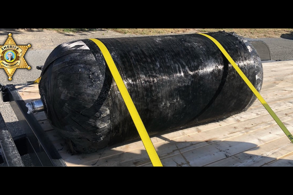 A photo of a large black cylinder strapped to a wood platform with yellow cords