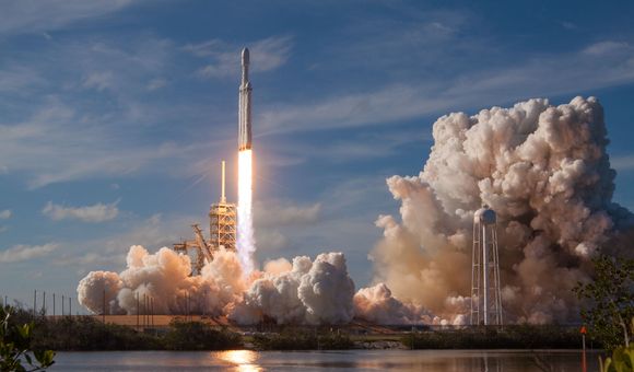 A rocket launches into a bright blue sky from a launchpad amid a great deal of smoke and near a body of water