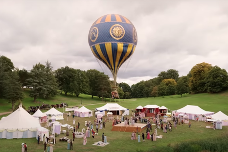 A vibrant hydrogen balloon with intricate blue and gold designs floats above a bustling outdoor event set against a cloudy sky. Below, several white tents are set up in a grassy park, with groups of people mingling and walking around.