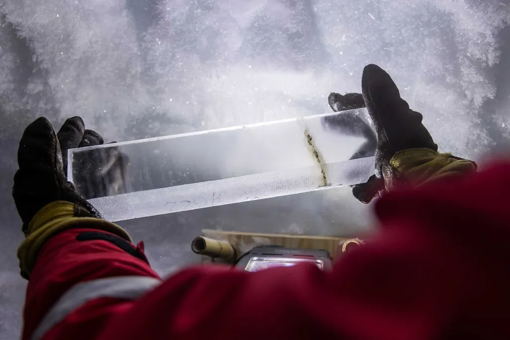 a person in a red jacket holds a roughly foot long ice core with black gloves above the snow