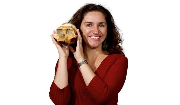 Headshot of paleoanthropologist Briana Pobiner in a red sweater holding an early human skull up text to her face on white background.