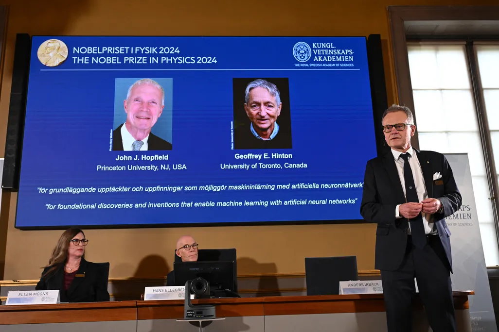 Man in a black suit standing in front of a projector screen with photos of two men