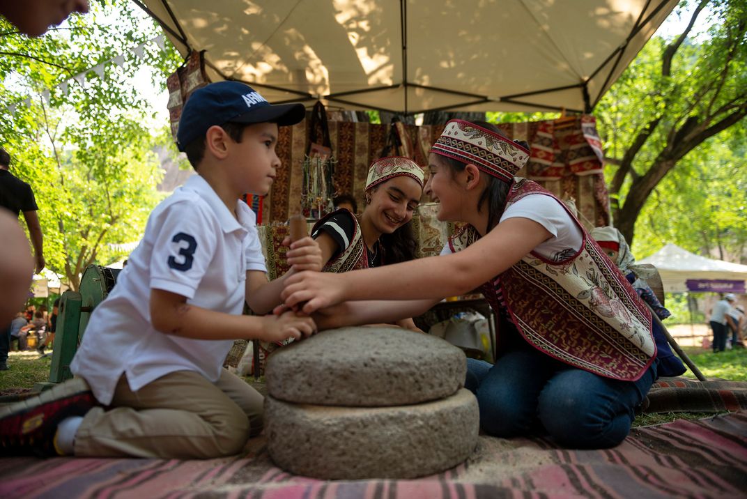 Two girls in Armenian dress and a young boy wearing a baseball cap sit together on the ground, smiling. They are seated around two stones placed on a rug, outdoors.
