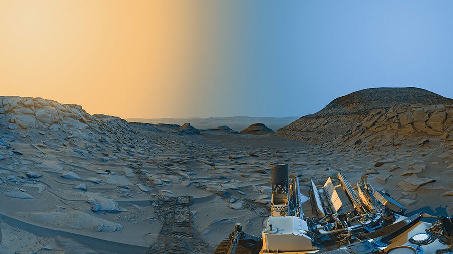 A stunning panorama of the Martian desert with a twilight sky. The landscape appears light rust and dull blue and is covered with boulders. A hill can be seen on the right.