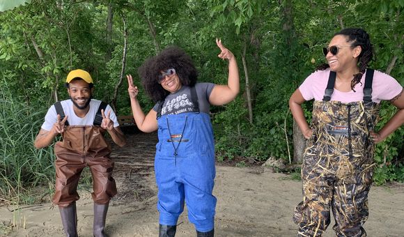 Three adults wearing chest waders laughing and in silly poses. They are standing on sand behind a line of plastic bins filled with water.