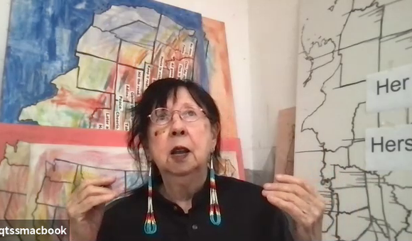Still from a video of Jaune Quick-to-See Smith. Smith wears a black shirt with her hair pulled back, gold wire glasses, and long beaded earrings. She is sitting in front of works of art depicting maps of the United States, several in color and one in blac