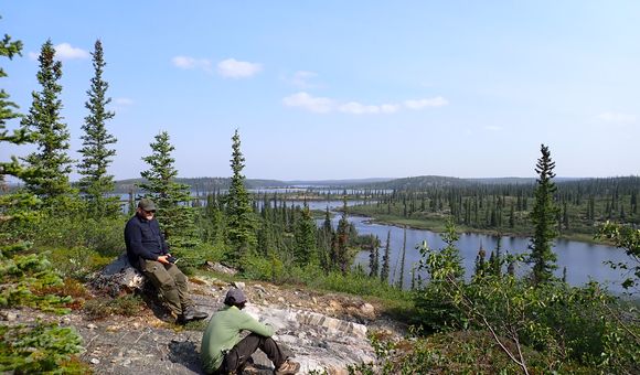 Two men sit on a rock bed on top of a forested hill over looking a remote lake.
