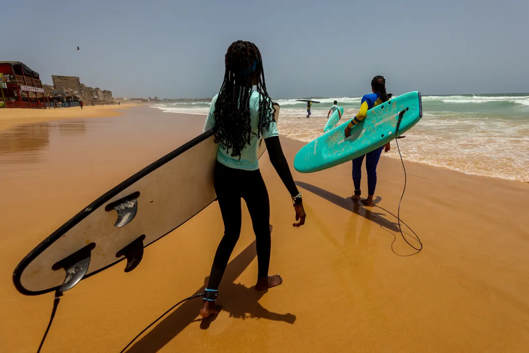 two girls walk on a beach carrying surfboards