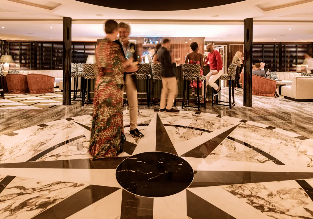 People gather around a stylish bar with a black and white marble compass design on the floor, socializing and enjoying drinks in an elegant lounge with sofas and armchairs in the background.