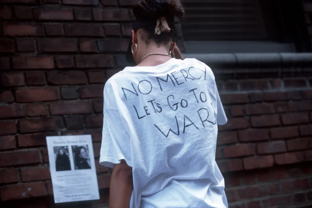 A woman faces away from the camera and looks at a missing person poster on Sept 13. She wears a white shirt with the handwritten message NO MERCY, LET'S GO TO WAR