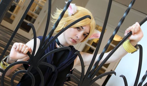 A person in a costume of black tank top, short yellow wig with furry white ears, and yellow leather bracelets, poses through a black wrought-iron railing.