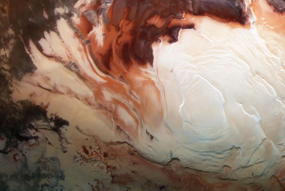 An image of Mars's South Pole. The photo shows a white icy cap surrounded by swirls of various shades of red. 