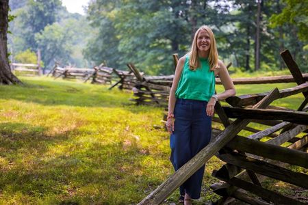 A woman leans on a snaking, split rail fence on a mossy green field.