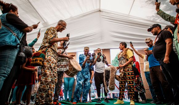 Four men stand together on a tented stage playing percussive instruments. They are dressed in brightly patterned clothes and are filmed by people holding cellphones.