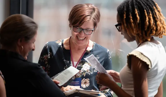 Three women stand in a circle laughing and discussing printed handouts