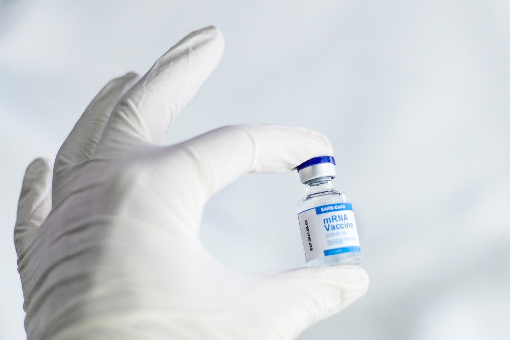 A white gloved left hand holds a small glass vial containing doses of the pfizer covid-19 vaccine.