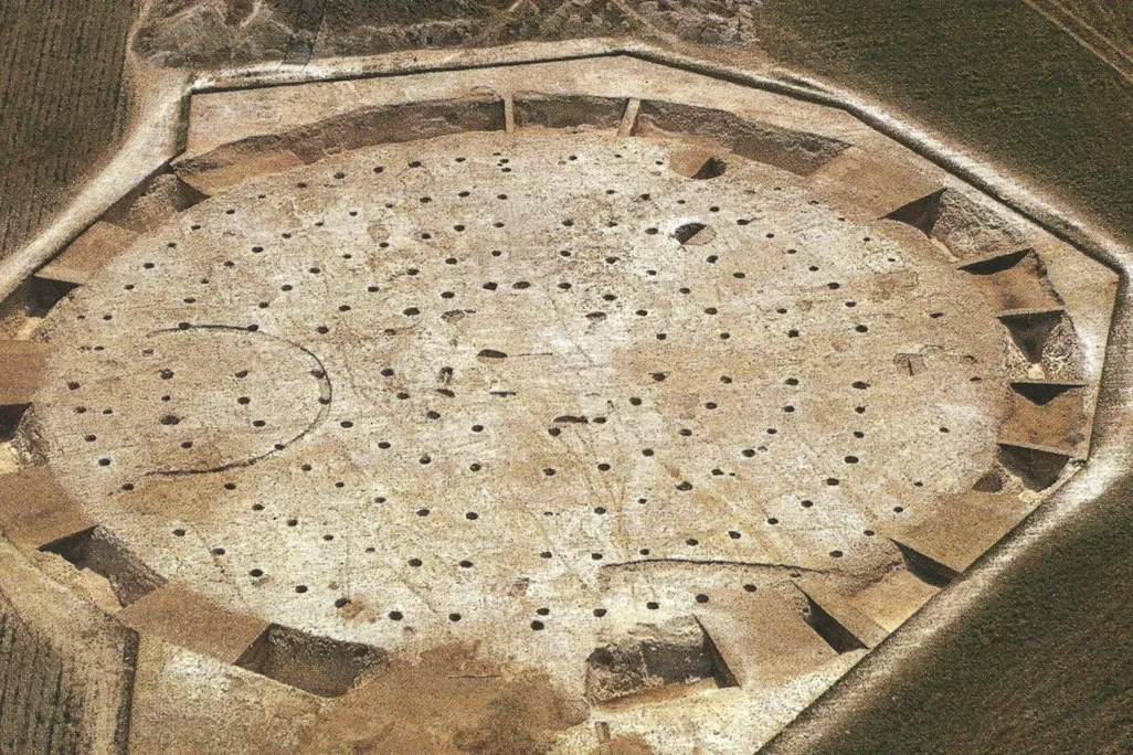 An aerial view of sandy dirt ground with numerous holes in concentric circles, and a ditch surrounding the entire formation
