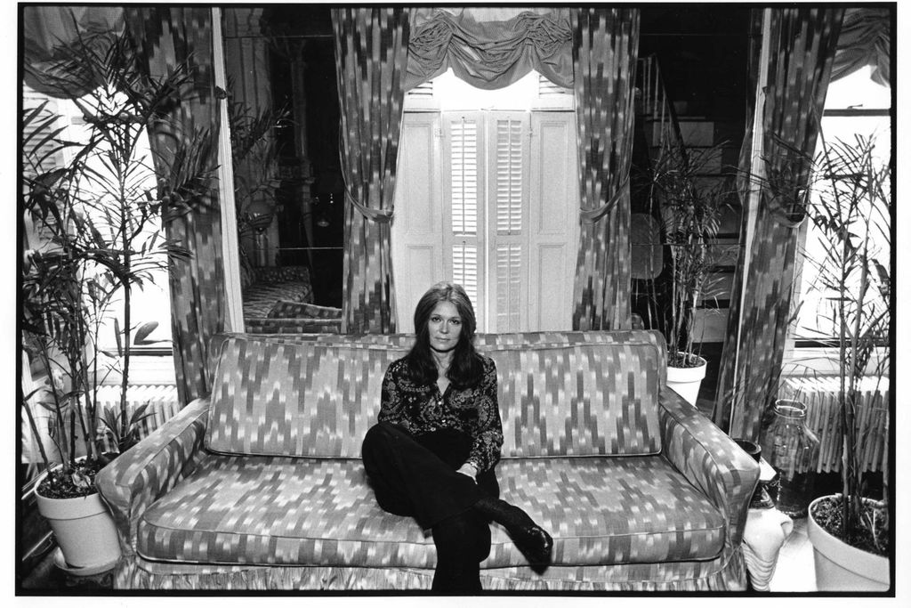 A black and white image of Steinem, a white woman in a patterned button down and black pants, sitting on a patterned couch in front of house plants and tall windows with curtains