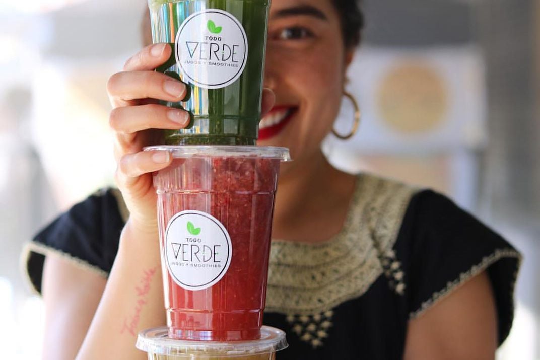 A smiling woman holds two agua frescas stacked on top of each other, in front of her face.