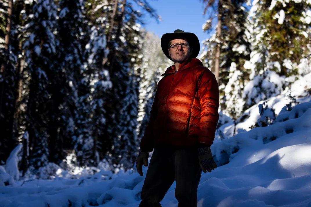 a man in a red winter coat stands for a portrait in a snow covered forest