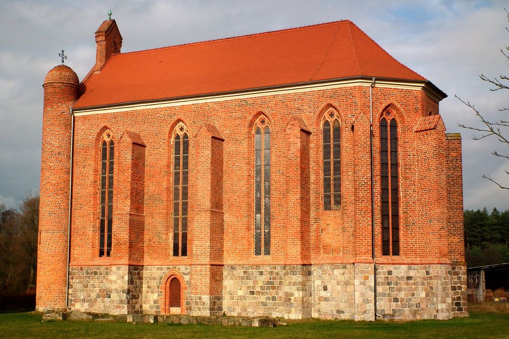 The exterior of an orange brick chapel with skinny, tall Gothic windows, a gabled red roof and a curved nave on one end.
