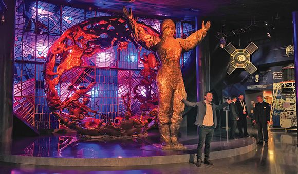 A museum display features a statue of a male astronaut standing with his arms outstretched in front of a metal sculpture lit by red and purple lighting. A museum visitor stands in a similar pose in front of the statue.