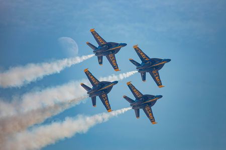 Four military jets fly in a diamond formation across a blue sky. On the underside of each jet's wings, "US Navy" is painted in bold yellow letters. White airshow smoke streams from each jet. The moon is visible in the sky.