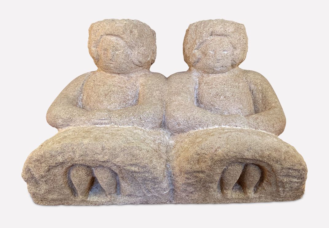 A view of a white-ish stone sculpture of two women, seated side by side with their small feet poking out of their identical dresses