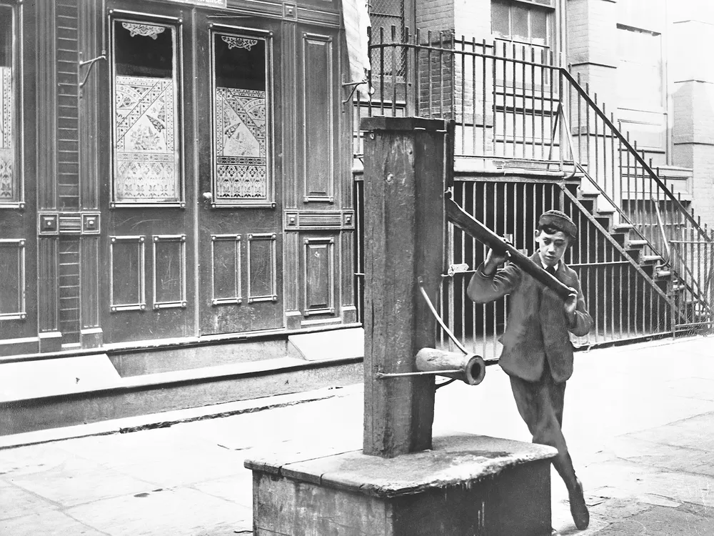 An old photo of a boy holding the handle of water pump on a street in New York City