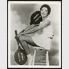 Althea Gibson poses on a stool with two tennis rackets. Both the rackets have dark colors printed with "Althea Gibson" in white letters.