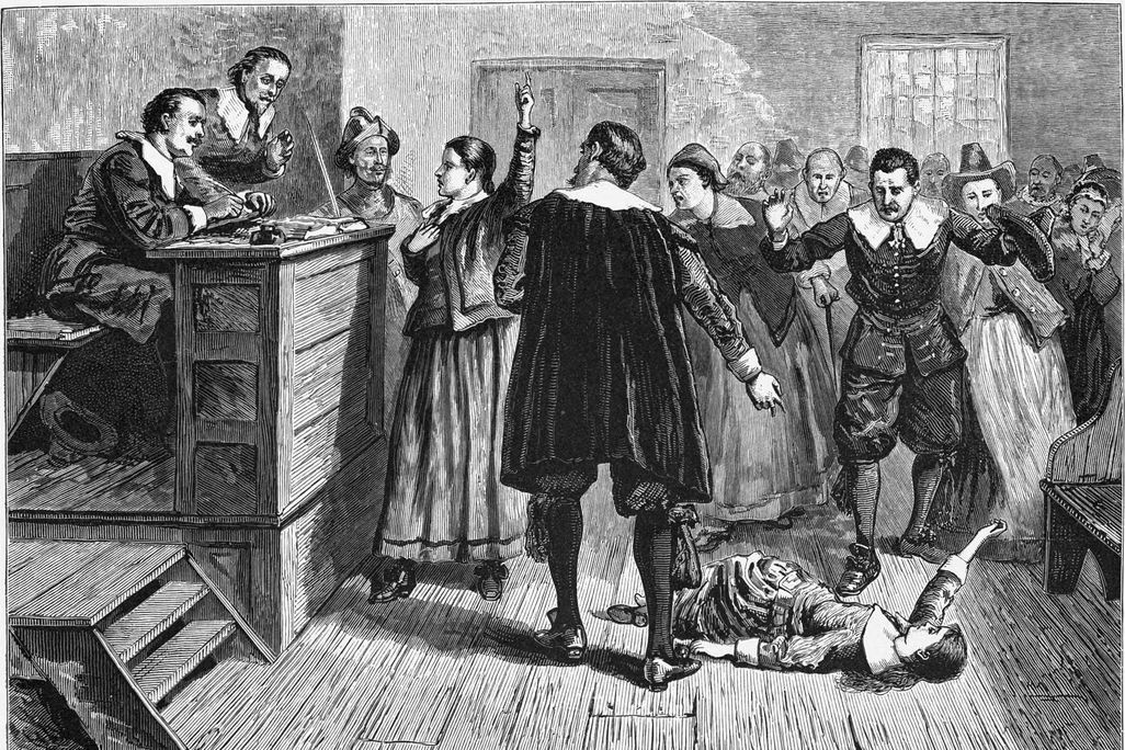 A black and white engraving of a courtroom scene; one woman stands in the center and raises a hand as though defending herself, while a young girl falls on the floor beneath a crowd of people