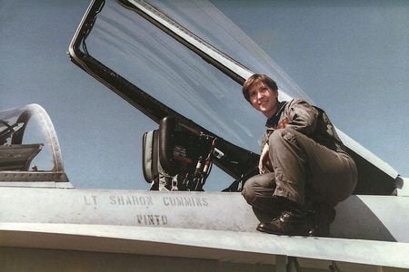 A young woman wearing a U.S. Navy olive-green flightsuit and black combat boots perches next to the cockpit of her F/A-18C miltary jet. "Lt Sharon Cummins" is stenciled onto the side of the airplane.