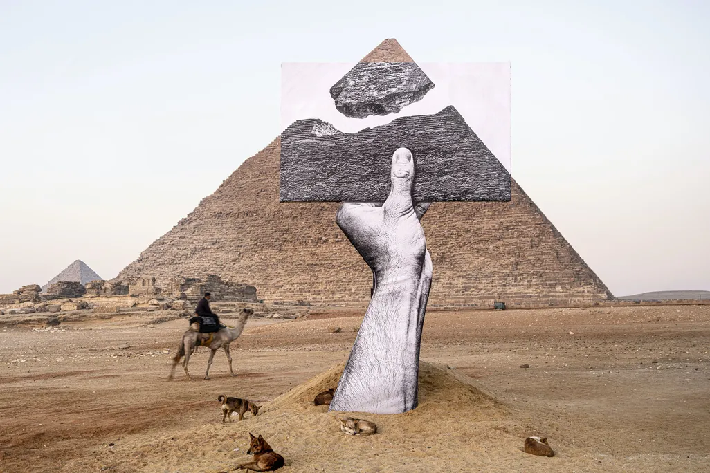A disembodied black and white hand emerges from the sand and holds a photograph, which creates the effect that the tip of a pyramid behind is separated and floating above its base