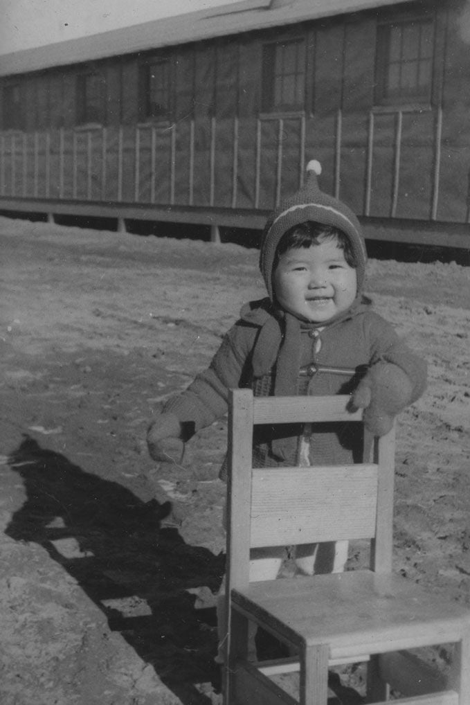 A black and white photo shows a small child smiling and holding a chair in front of a chainlink fence.